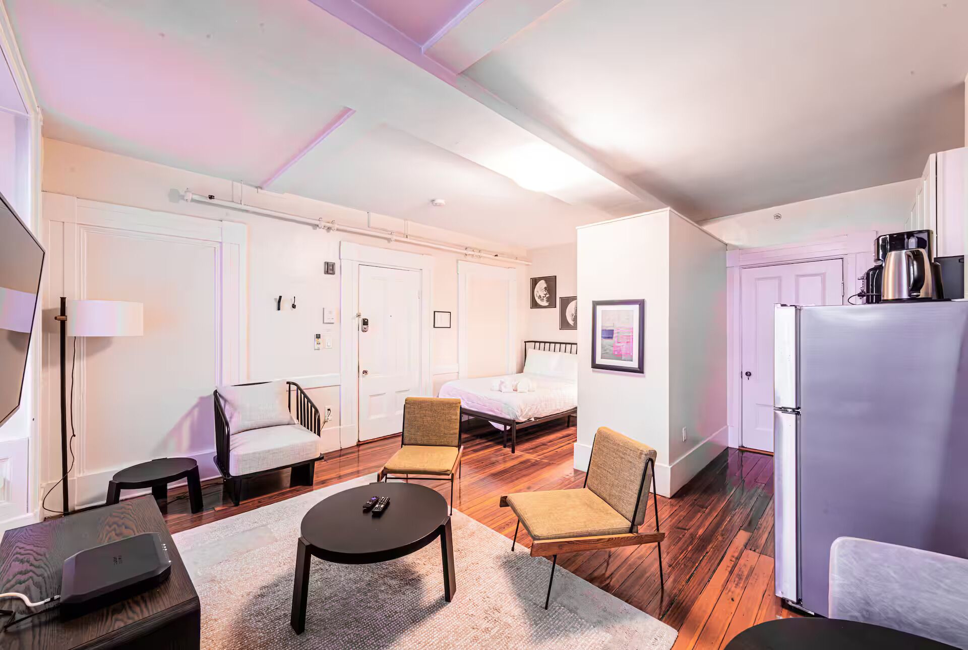 A compact room with a bed, two chairs, a small table, a refrigerator, and a lamp.