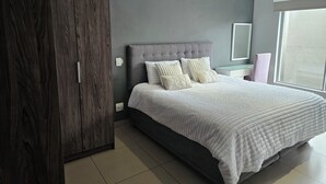 Une chambre à coucher avec une tête de lit rembourrée de couleur grise, un grand lit avec une literie blanche, une armoire en bois et une fenêtre donnant sur un beau paysage.