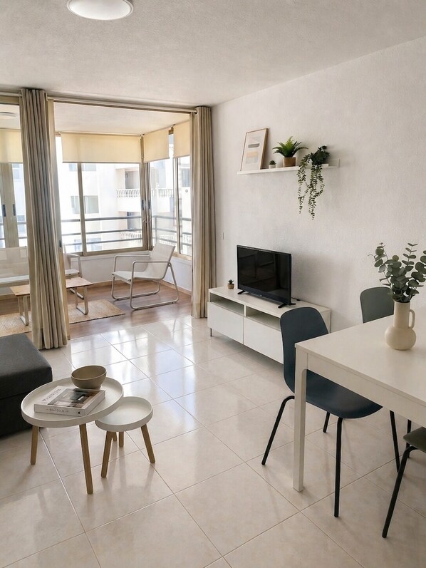 Un salon moderne avec un téléviseur à écran plat, une table à manger et des chaises. On y trouve un canapé, une petite table basse et une plante en pot sur le rebord de la fenêtre.