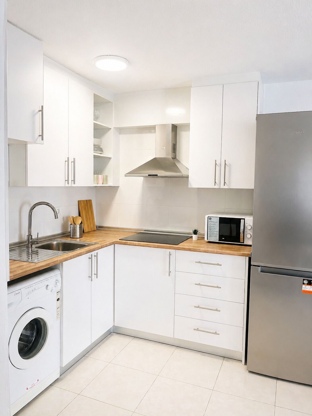 Een compacte keuken met witte kasten, een roestvrijstalen koelkast, een magnetron, een spoelbak en een wasmachine.