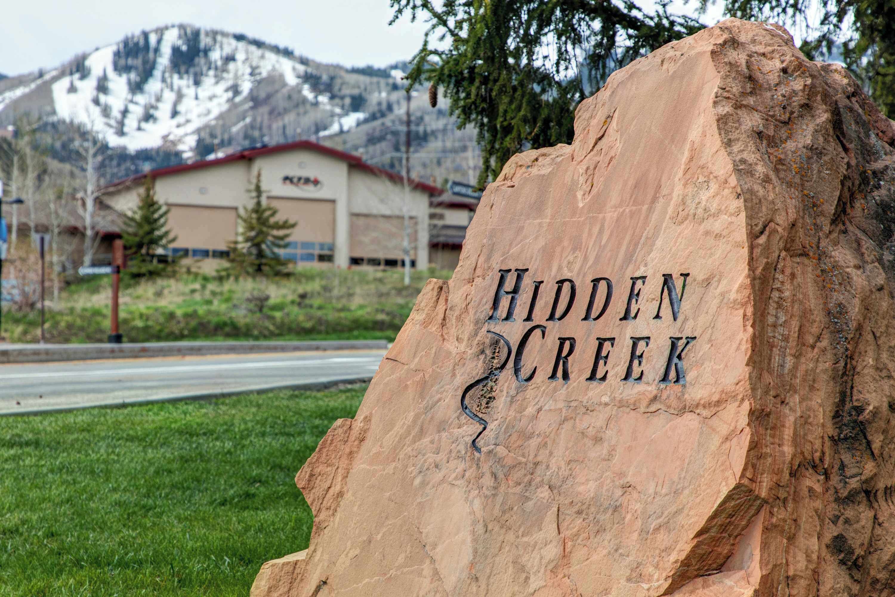 A large rock with 'Hidden Creek' engraved on it, a building with a red roof in the background, and a mountain with snow.