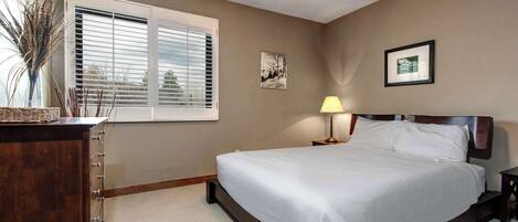 A bedroom with a bed, a bedside table, a lamp, a window with blinds, and a wall-mounted picture.