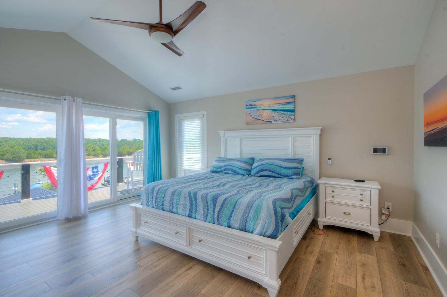 A bedroom with a bed, a nightstand, a ceiling fan, a painting, and a view of the outdoors.