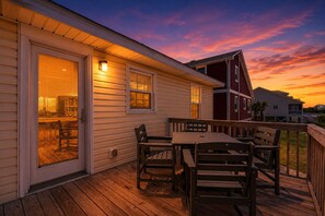 Une terrasse en bois avec une table et des chaises, une porte d’entrée en verre et une vue sur le coucher de soleil.