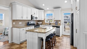 Une cuisine avec des meubles blancs, des plans de travail en granit, un îlot central avec des tabourets, un ventilateur de plafond et une vue sur l’extérieur, visible à travers la porte.