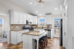A kitchen with white cabinets, granite countertops, a central island with stools, a ceiling fan, and a view through the door. - Shout! at the Shore: 4BR/2BA , Ocean Views, perfect for families! (Oak Island)