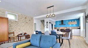 Ein modernes Wohnzimmer mit Steinwand, einem blauen Sofa, einem Essbereich mit Tisch und Stühlen sowie einer Küche mit blauen Wänden und weißen Schränken.