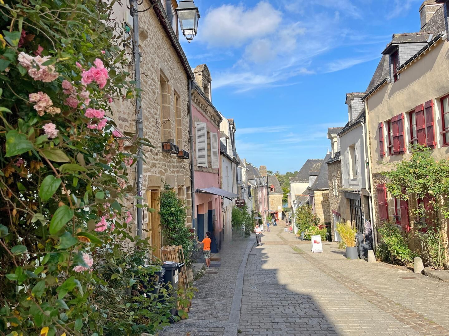 A cobblestone street lined with historic stone buildings, some with shutters and signage, under a clear blue sky.