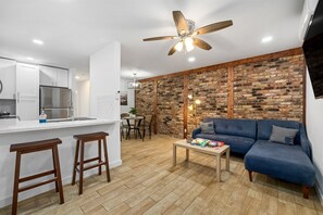 A modern kitchen with white cabinets, a stainless steel refrigerator, and a wooden bar stool. There is a blue sofa and a wooden coffee table in the living area. - Pet-Friendly Montrose 1BR | Museums| Free Parking newly renovated M2 (Houston)