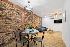 A dining area with a brick wall, a round table with chairs, a ceiling fan, and a flat-screen TV mounted on the wall. - Pet-Friendly Montrose 1BR | Museums| Free Parking newly renovated M2 (Houston)