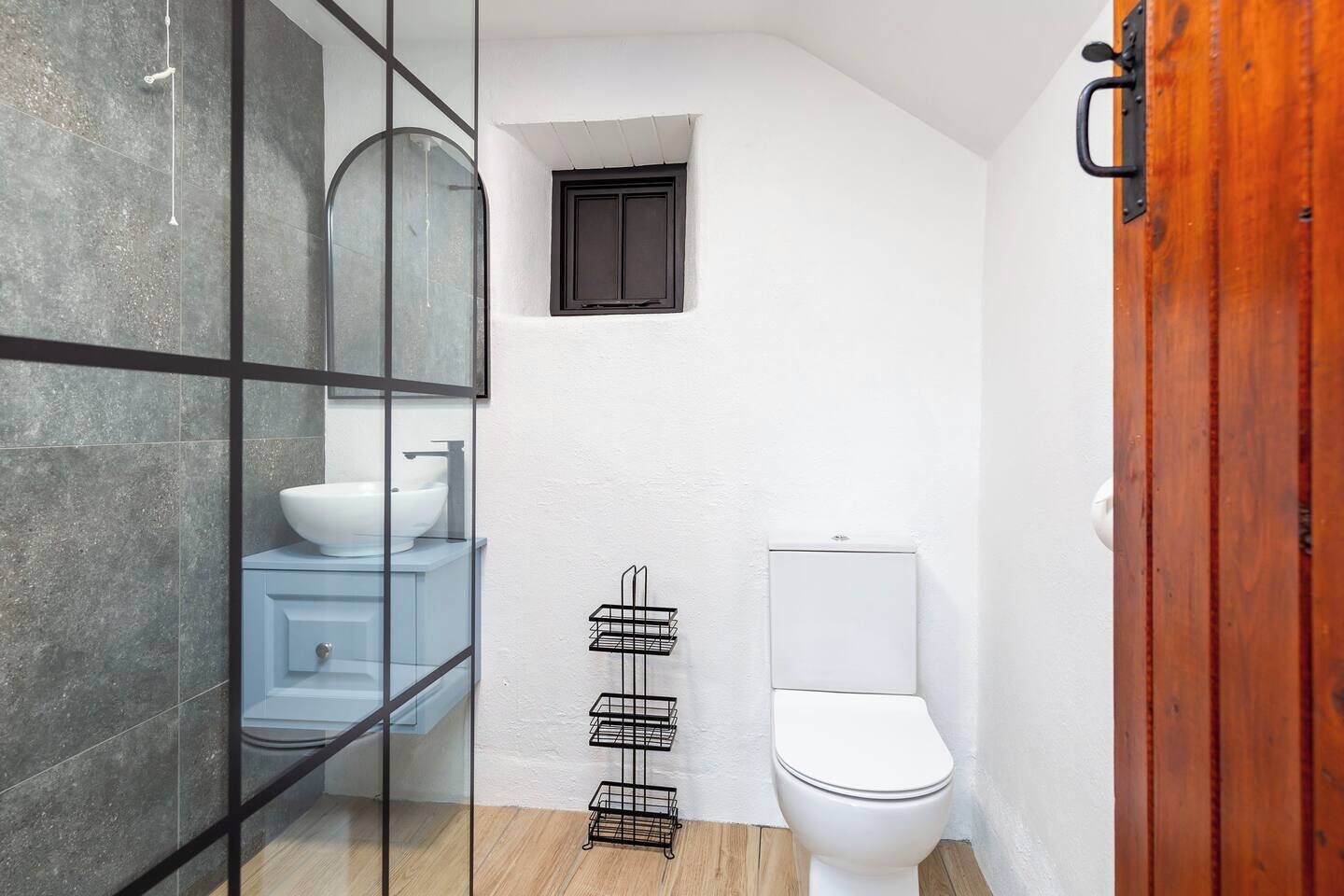 Une salle de bain avec une cloison en verre, des toilettes blanches, un meuble-lavabo bleu et une petite fenêtre.