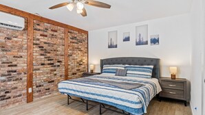 Een slaapkamer met een bakstenen muur, een bed met een gestreept dekbed, een plafondventilator en aan de muur bevestigde fotolijstjes.