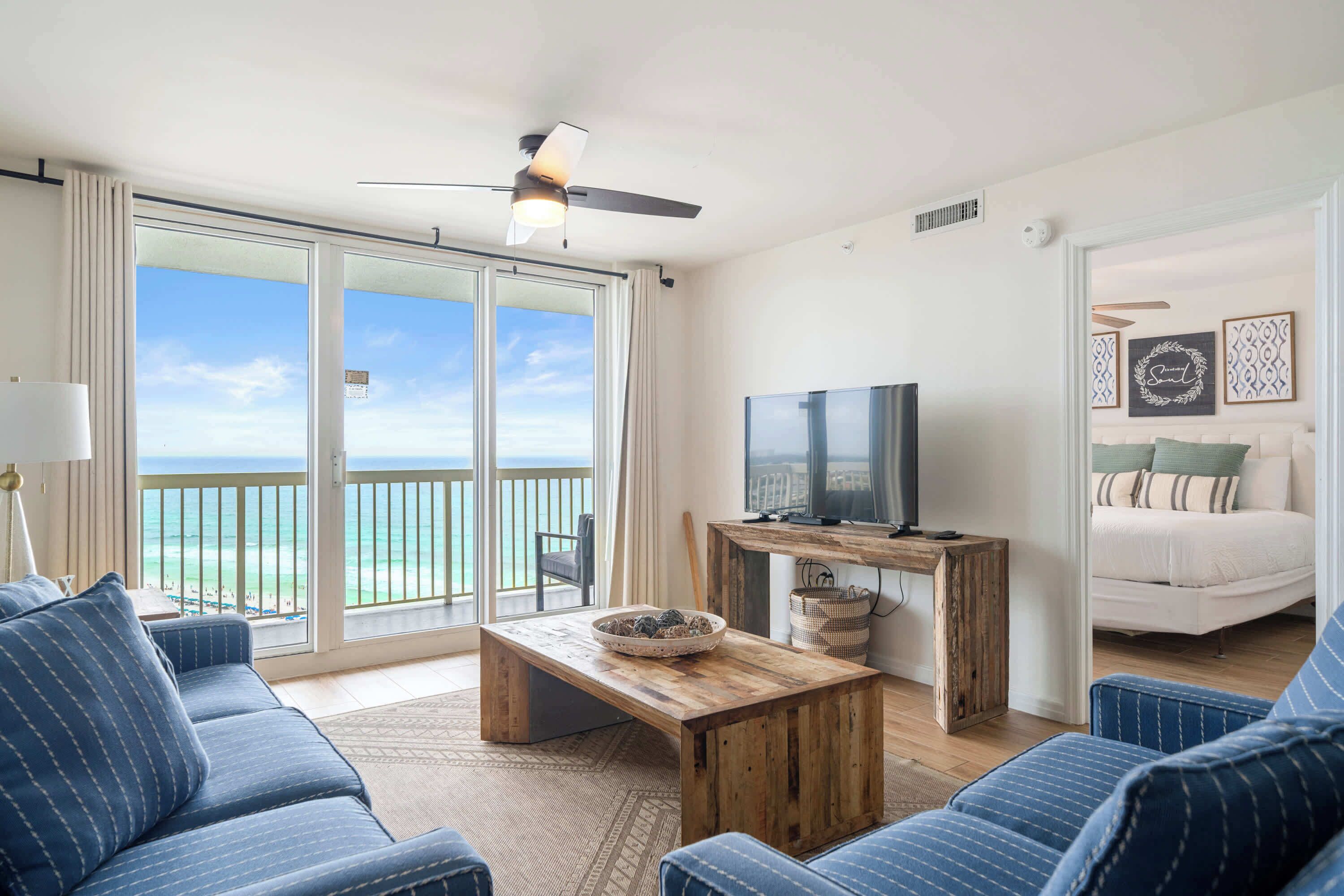 A room with a large sliding glass door leading to a balcony, a television on a wooden stand, a sofa, and a bed with a view.