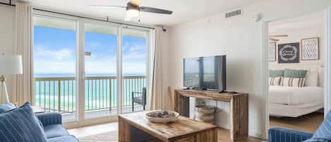 A room with a large sliding glass door leading to a balcony, a television on a wooden stand, a sofa, and a bed with a view.