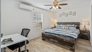 Ein Schlafzimmer mit einem hölzernen Bett, einem Schreibtisch und einem Stuhl. Es gibt einen Deckenventilator, ein Fenster mit Jalousien und Wandbilder.
