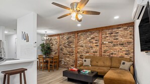 Una sala moderna con pared de ladrillo, un sofá marrón, una mesa de centro y un comedor con encimera blanca y sillas de madera.