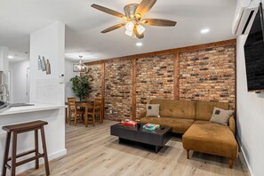 A modern living room with a brick wall, a brown sofa, a coffee table, and a dining area with a white countertop and wooden chairs. - Pet-Friendly Montrose 1BR | Museum District | Free Parking<br>newly renovated (Houston)