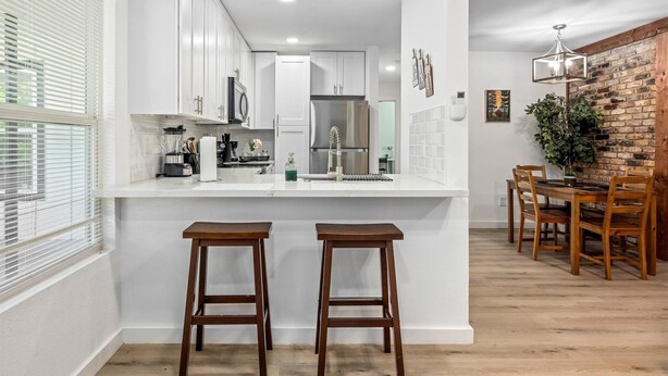 Una cocina moderna con armarios blancos, un refrigerador de acero inoxidable y un comedor con sillas de madera y una pared de ladrillo.