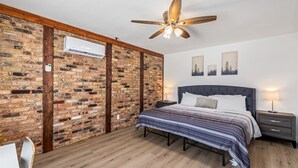 Ein Schlafzimmer mit Backsteinwand, einem Bett mit gestreifter Bettdecke, einem Nachttisch mit Lampe und einem Deckenventilator.