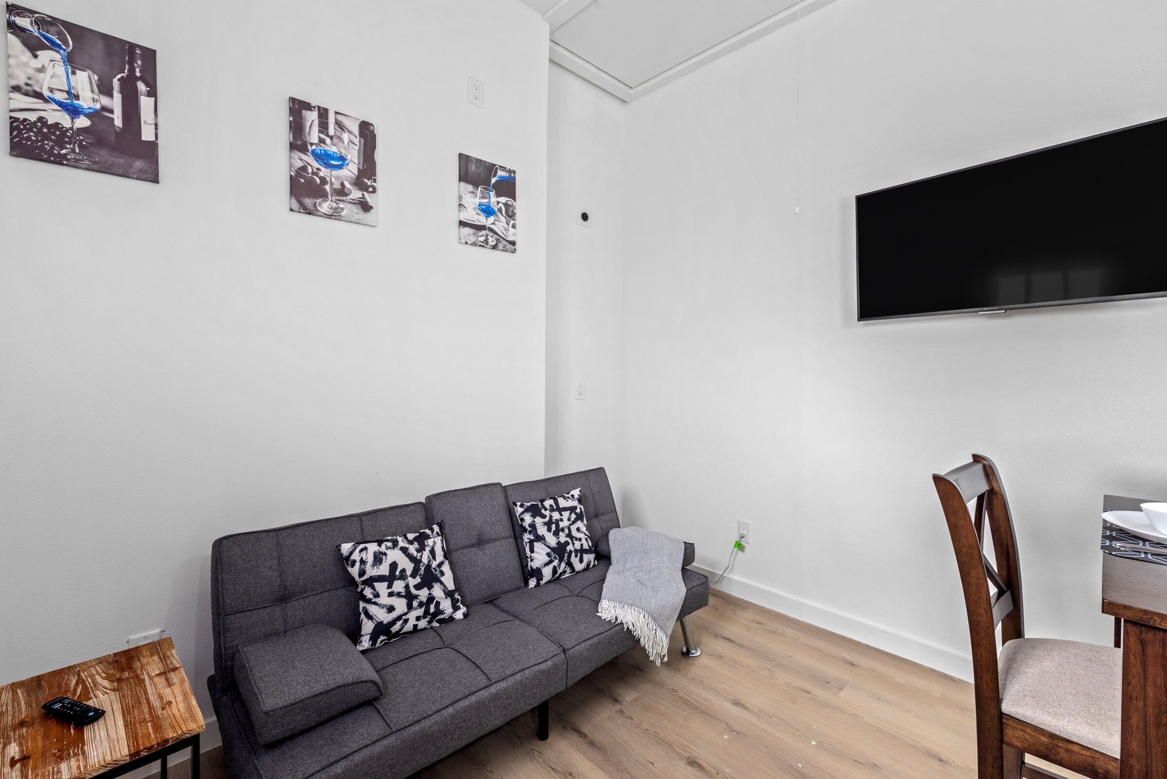 Un salon moderne avec un canapé gris, une table d’appoint en bois, un téléviseur fixé au mur et un coin repas avec une chaise en bois.