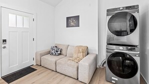 Ein moderner Waschraum mit einer Frontlader-Waschmaschine und einem Trockner, ein beiges Sofa mit Überwurfdecke und ein kleines gerahmtes Bild an der Wand.