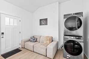 Ein moderner Waschraum mit einer Frontlader-Waschmaschine und einem Trockner, ein beiges Sofa mit Überwurfdecke und ein kleines gerahmtes Bild an der Wand.