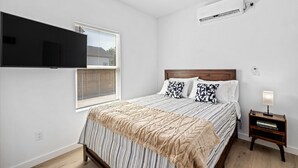 Ein Schlafzimmer mit einem Bett, einem an der Wand montierten Fernseher, einem Deckenventilator, einer Klimaanlage, einem Nachttisch mit Lampe und einem Fenster mit Jalousien.
