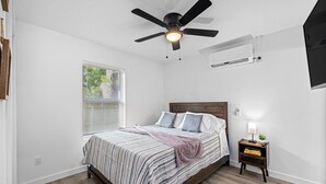 Een slaapkamer met een bed, een nachtkastje, een plafondventilator, een raam met jaloezieën en een airconditioning.