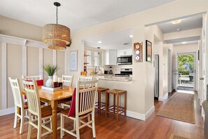 A dining area with a wooden table and chairs, a hanging light fixture, and a kitchen with white cabinets and appliances. - Spacious Recently Updated Bayside 4 Bedroom (Ocean City)