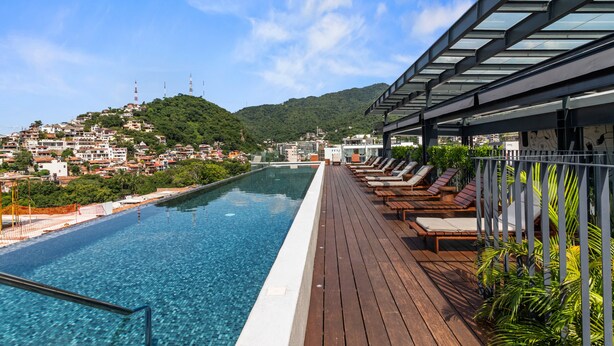 Zona junto a la piscina con cubierta de madera y sillas de descanso, con vistas a un paisaje urbano y colinas.