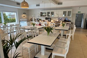 Un comedor moderno con mesas y sillas blancas, una cocina al fondo y vistas al exterior.