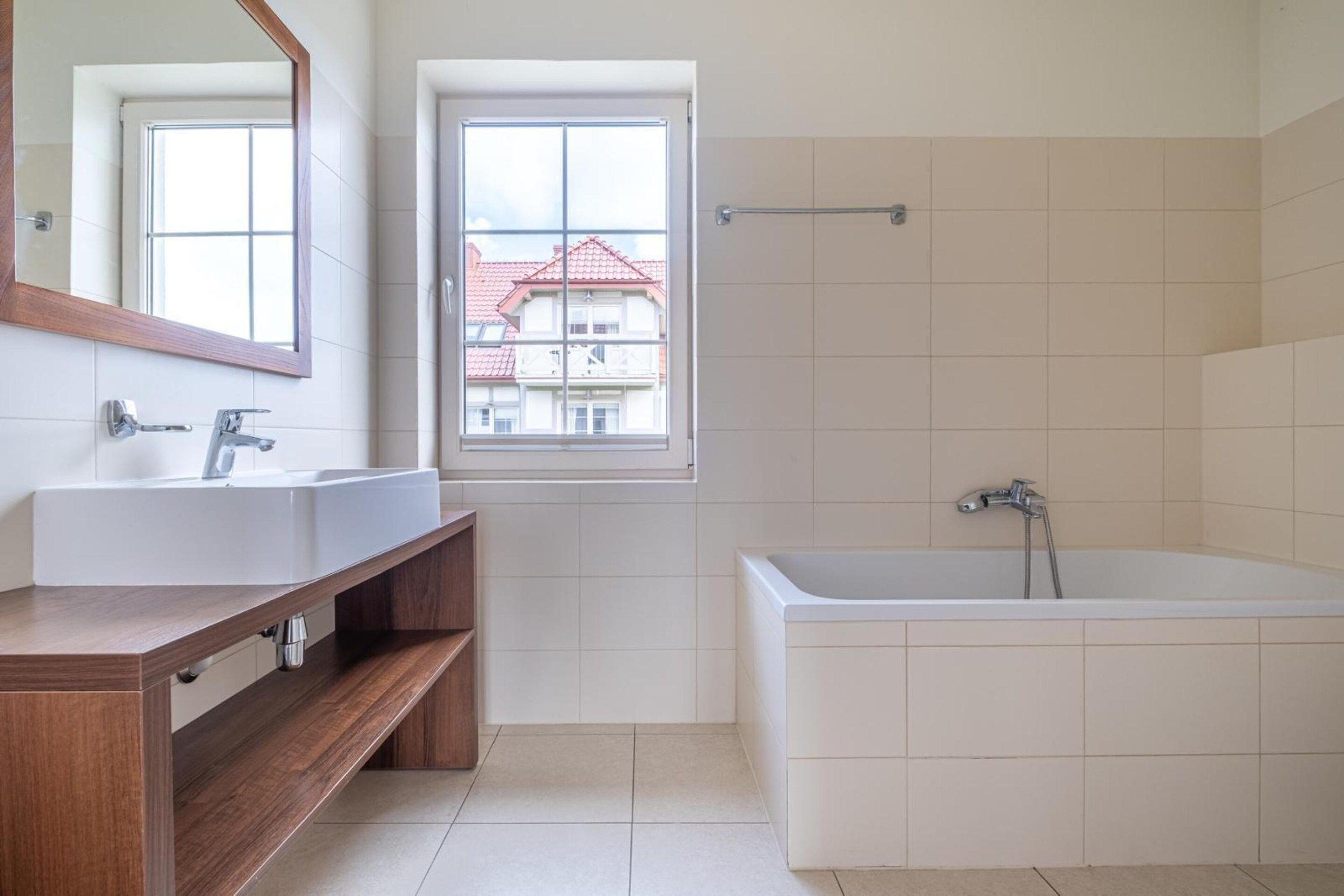 Een badkamer met een ruime badkuip, twee wastafels, een houten wastafelmeubel, een raam en uitzicht op gebouwen.