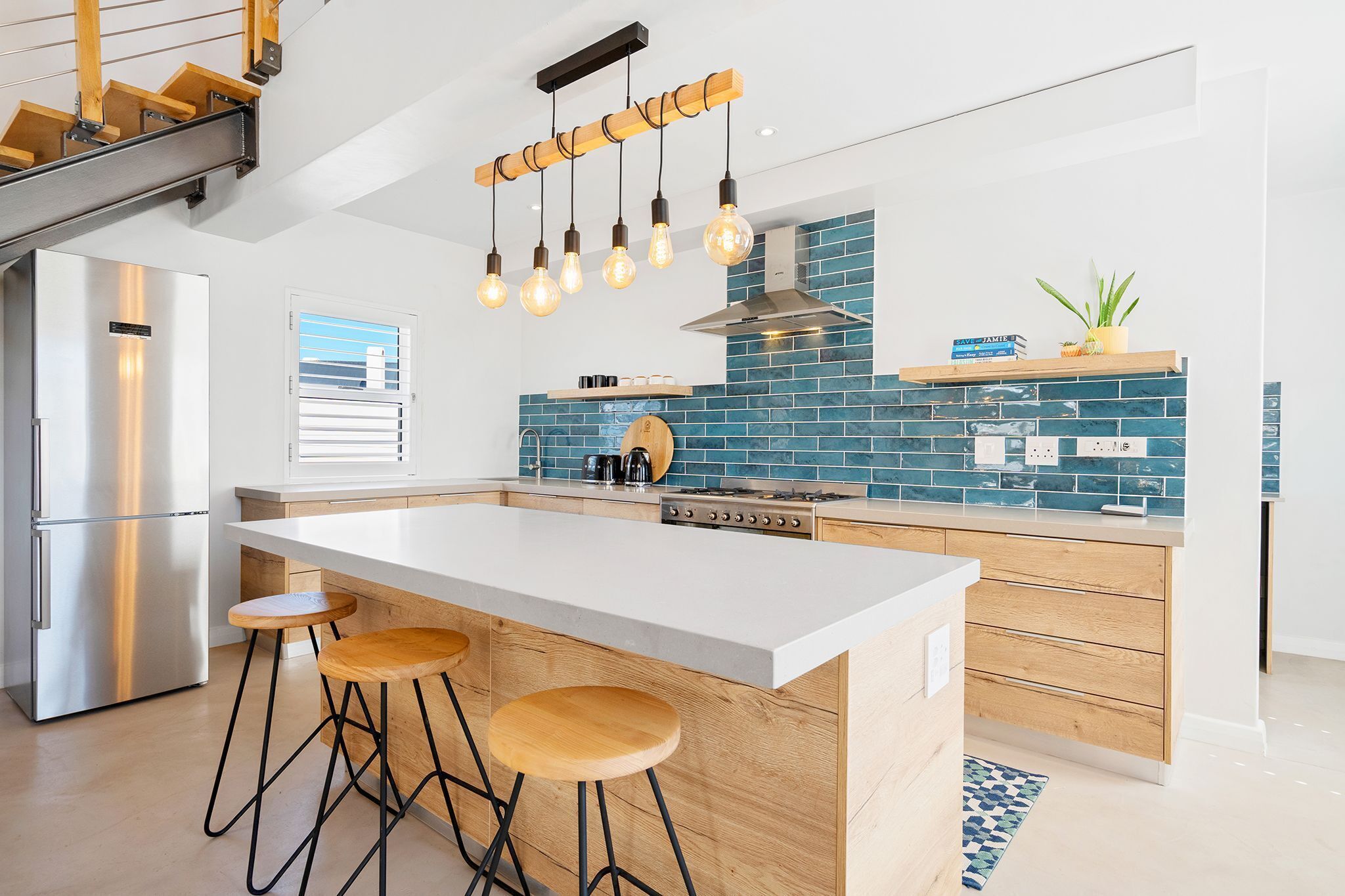 A modern kitchen with a central island, bar stools, a stainless steel refrigerator, and a blue-tiled backsplash.