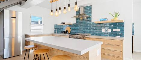 A modern kitchen with a central island, bar stools, a stainless steel refrigerator, and a blue-tiled backsplash.