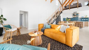 Un espacio habitable amplio con un sofá amarillo, sillas de madera, una escalera y una cocina al fondo.