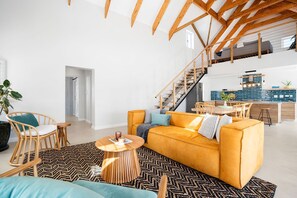 Uma sala de estar ampla com um sofá amarelo, cadeiras de madeira, uma escada e uma cozinha ao fundo.