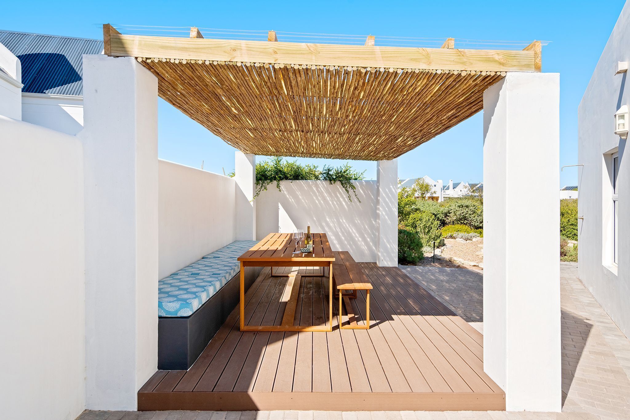 A wooden deck with a table and bench, a pergola overhead, and a white wall with a window.