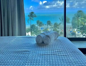 Ein Bett mit einer blauen, strukturierten Decke, ein Fenster mit Blick auf den Strand und zwei aufgerollte Handtücher.