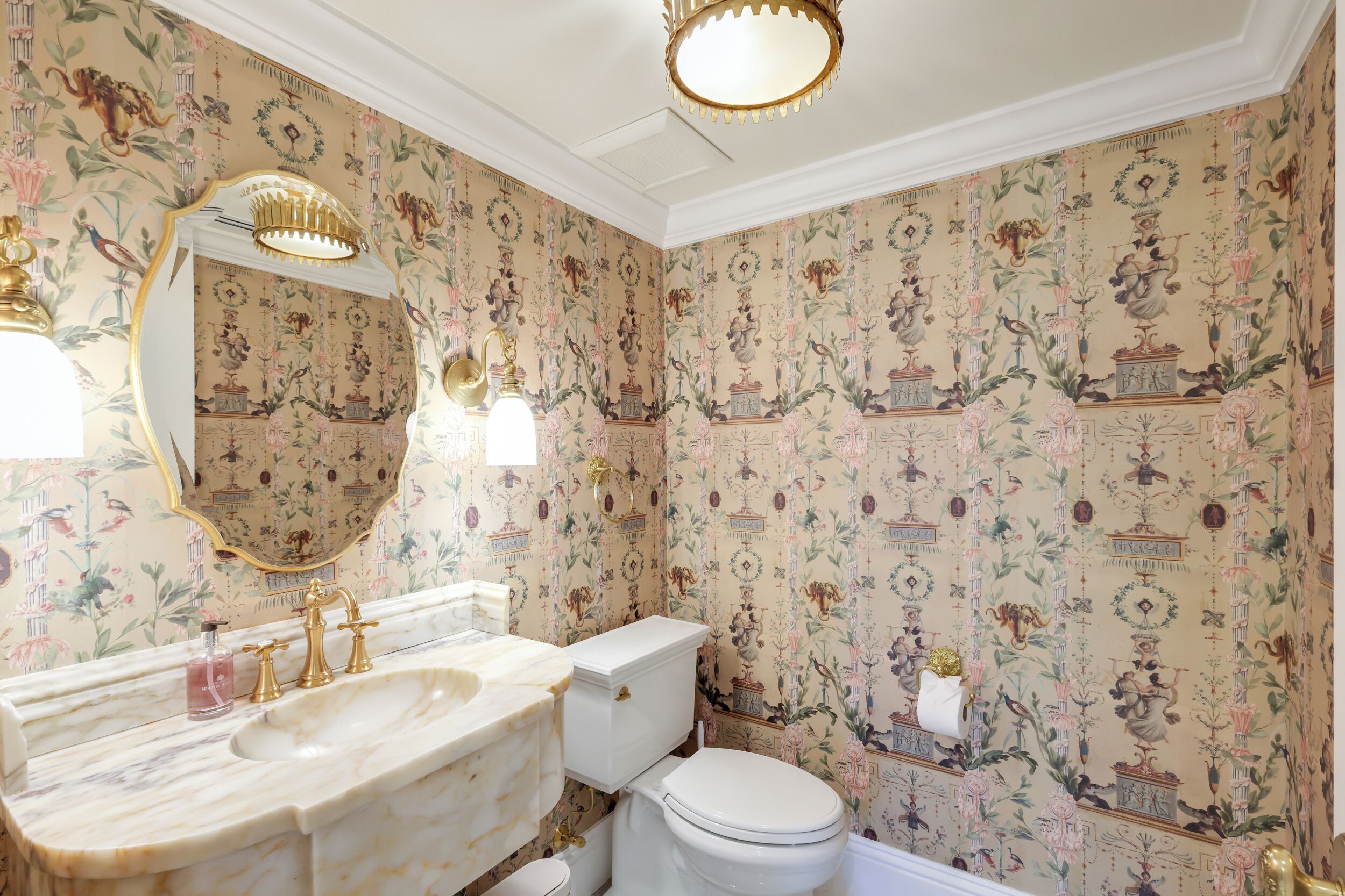 A bathroom with a marble sink, gold fixtures, a white toilet, and floral wallpaper.
