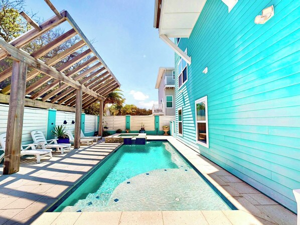 Una piscina con un porche de madera, sillas de descanso y una casa azul.