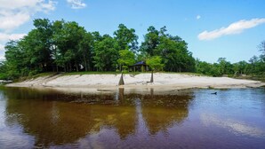 A riverside view with a sandy beach, trees, and a small pavilion.