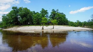 A riverside view with a sandy beach, trees, and a small pavilion.