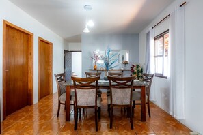 A dining area with a round table, six chairs, a mirror above the table, and a window with curtains. - Casa com Churrasqueira e Rede a 210 m da Praia (Navegantes)