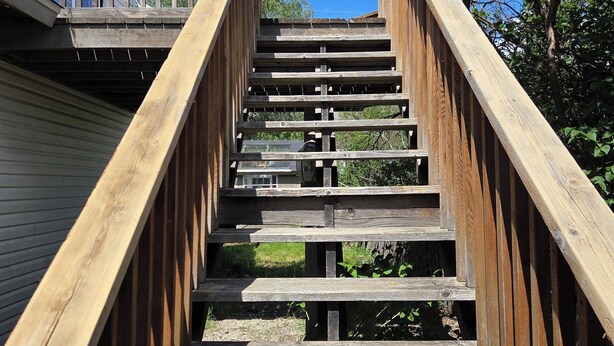 A wooden staircase leading to a deck with railings, surrounded by greenery.