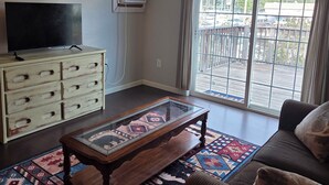 A living room with a television, a glass-top coffee table, a sofa, a patterned rug, a white dresser, and a sliding glass door leading to an outdoor deck.