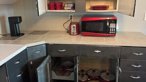 A compact kitchen with a microwave, toaster, and coffee maker on the countertop, and a cabinet with red mugs and plates.