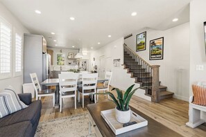 A modern kitchen with white cabinets, a dining area with a white table and chairs, a staircase with wooden steps, and a living room with a sofa and a coffee table.