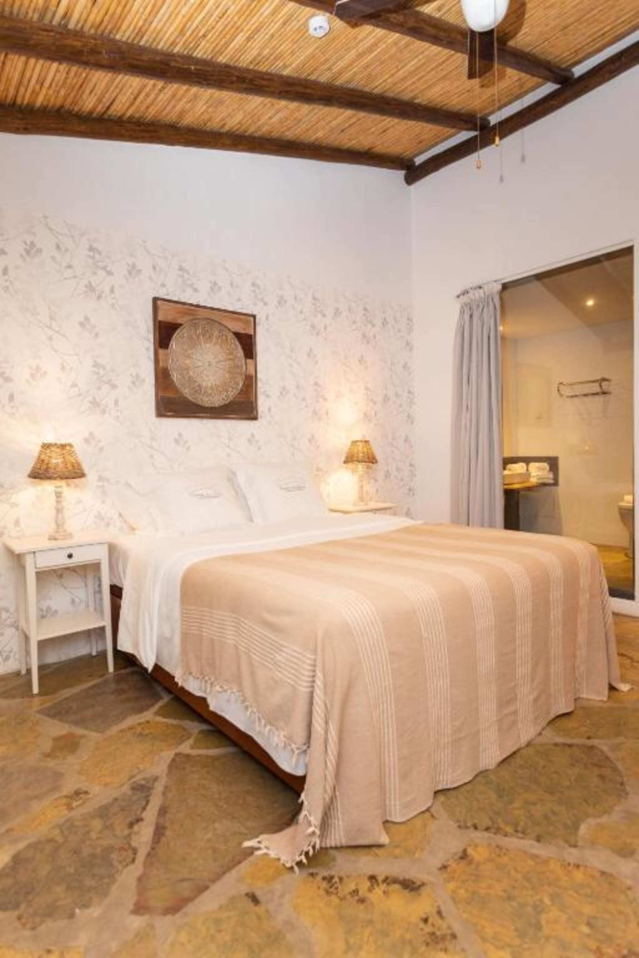 Un dormitorio con piso de piedra, una cama con cubrecama color beige, dos lámparas de noche y un plato decorativo en la pared.
