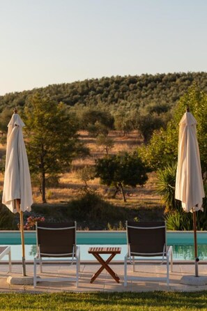 A poolside area with two lounge chairs and umbrellas, overlooking a hilly landscape.