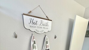 Un baño con un letrero que dice 'Baño Caliente LAVAR SUMERGIRSE RELAJARSE' y dos toallas a rayas colgadas de ganchos.